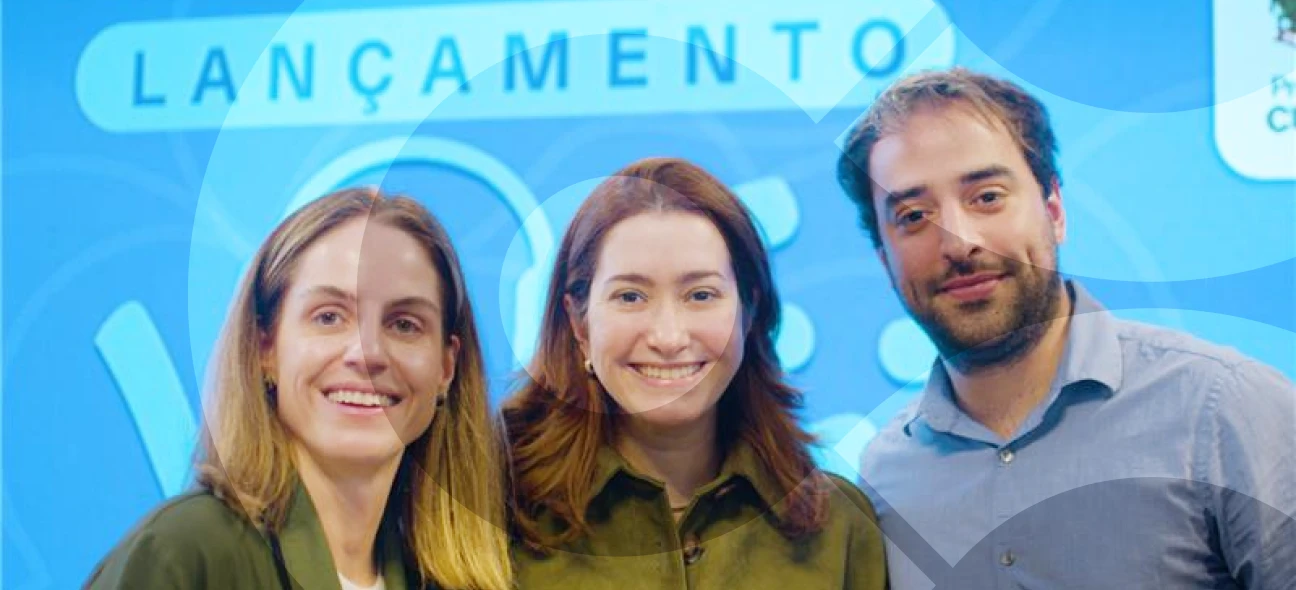 Três pessoas sorriem para a câmera em um evento de lançamento com fundo azul claro e o texto "LANÇAMENTO" ao fundo. O grupo é composto por duas mulheres, uma com cabelos loiros e outra com cabelos ruivos, ambas vestindo camisas verdes, e um homem com barba e cabelos curtos, vestindo camisa azul.