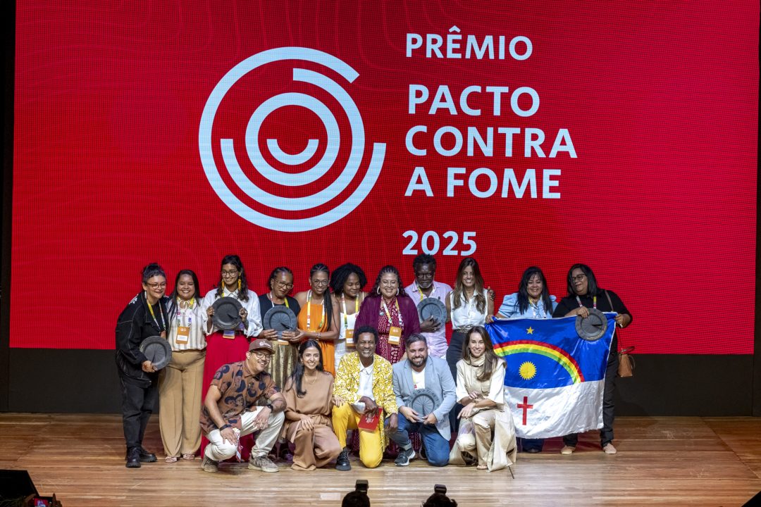 Premiados do Prêmio Pacto Contra a Fome 2025 reunidos no palco durante a cerimônia Grupo de premiados posa no palco em frente a um telão vermelho com o logo e o título “Prêmio Pacto Contra a Fome 2025”