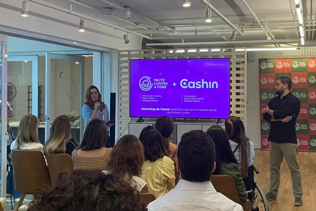 Apresentação da parceria entre Pacto Contra a Fome e Cashin durante evento corporativo Apresentação sobre a parceria entre Pacto Contra a Fome e Cashin, com a Luciana Ramos falando ao microfone diante de uma plateia e um telão exibindo o conteúdo da palestra.